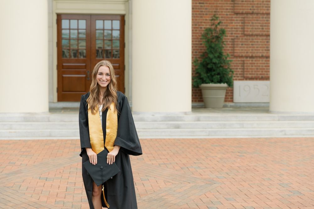Graduate in academic regalia poses in front of brick building with white columns.