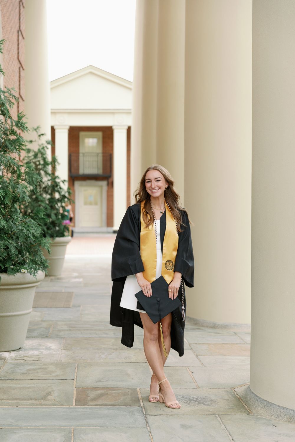 Graduate in black gown and gold stole sits between white columns on campus.