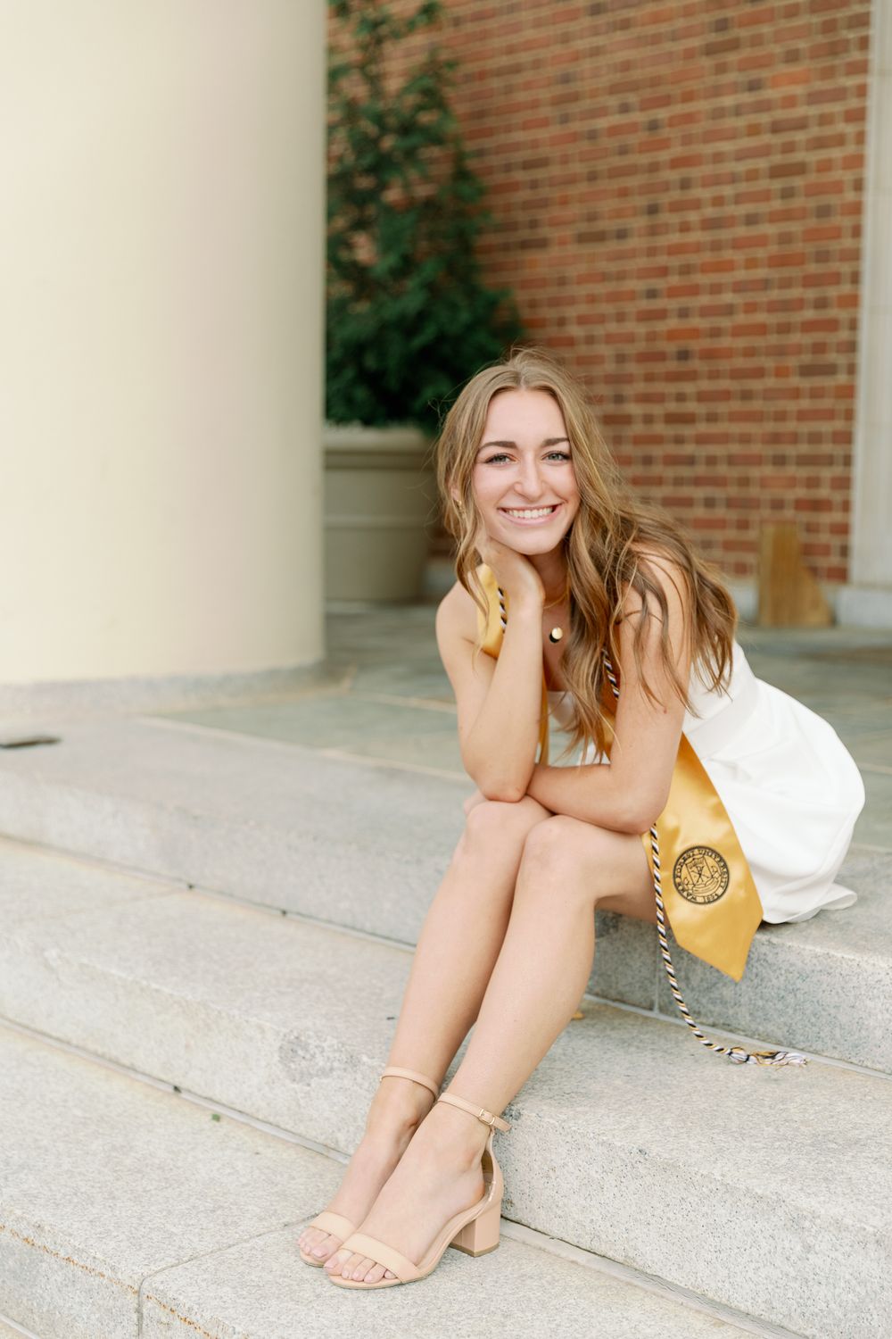 Graduate in white dress and gold stole poses casually on stone steps.