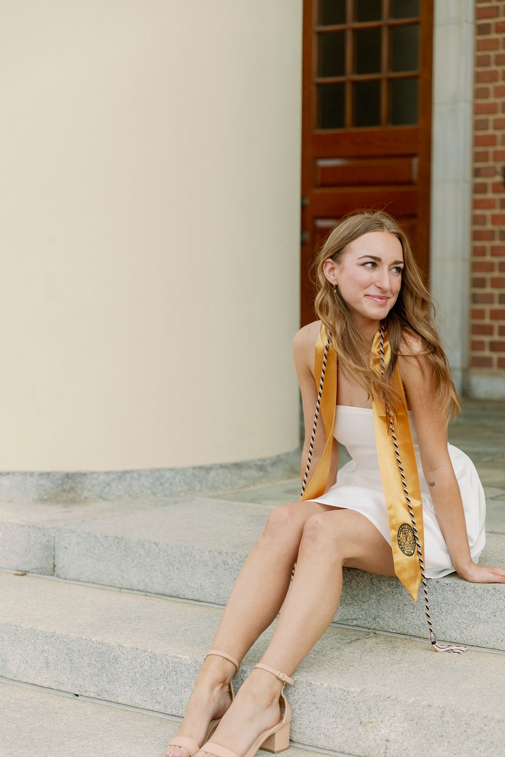 Graduate in white dress with gold honors stole relaxes on campus steps.