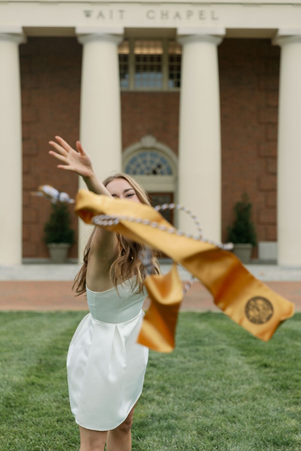 Graduate in white dress tosses gold stole in front of Wait Chapel.