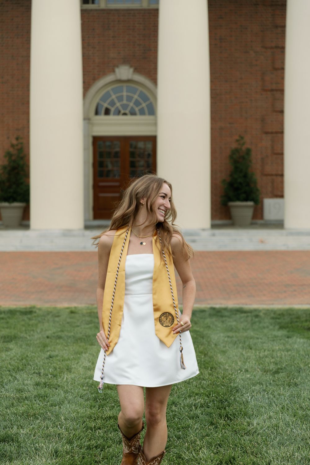 Graduate in white dress runs across lawn in front of columned building.