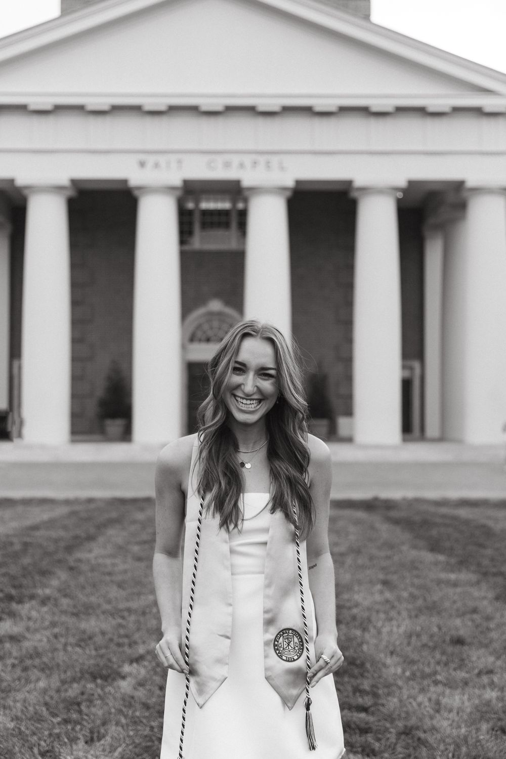 Graduate in white dress poses in front of columned building entrance with bright smile.