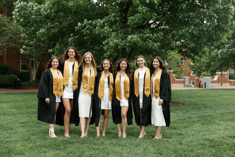 Group of graduates in black gowns and gold stoles pose together on campus lawn.