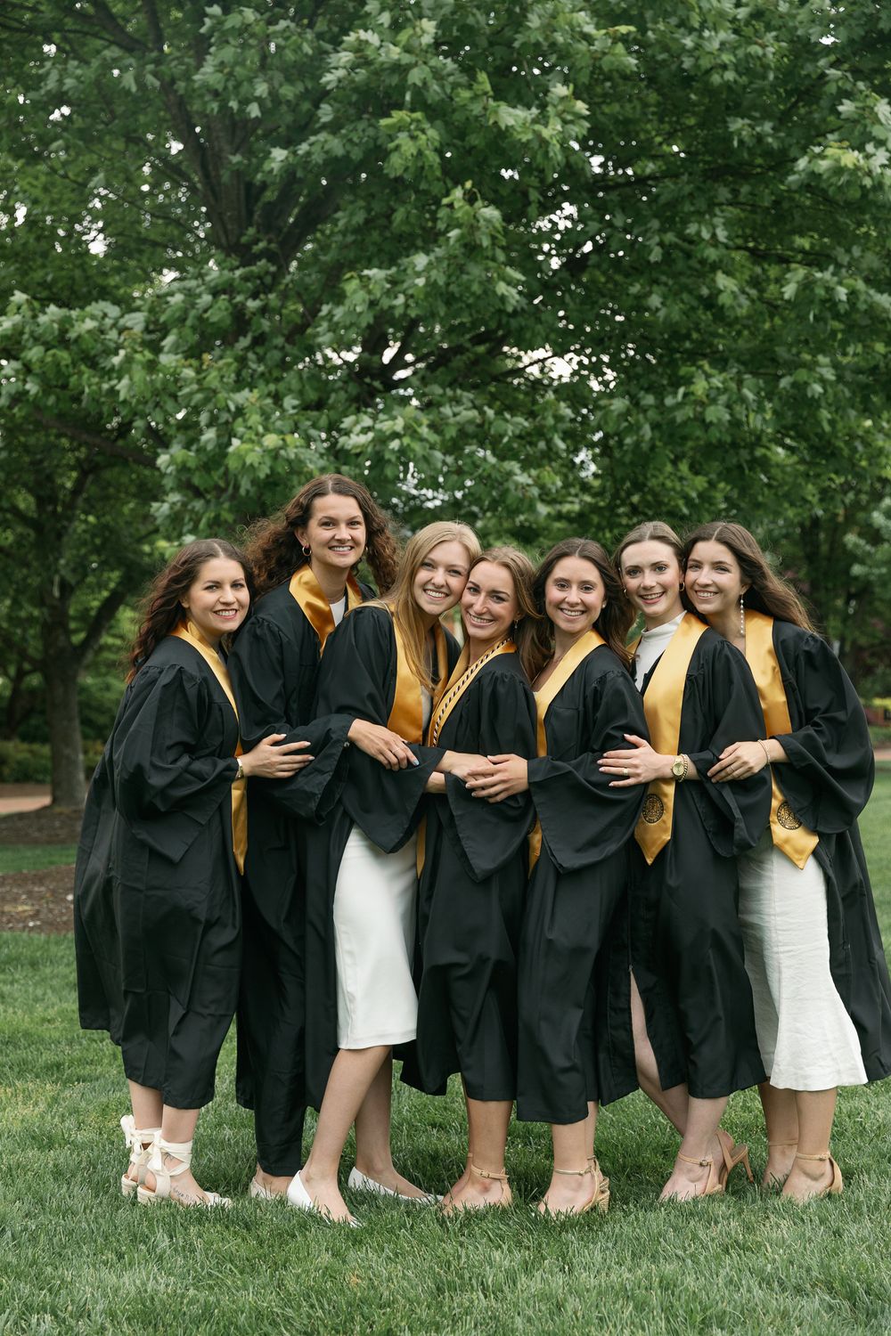 Seven graduates in black robes and gold stoles share candid moment on campus lawn.
