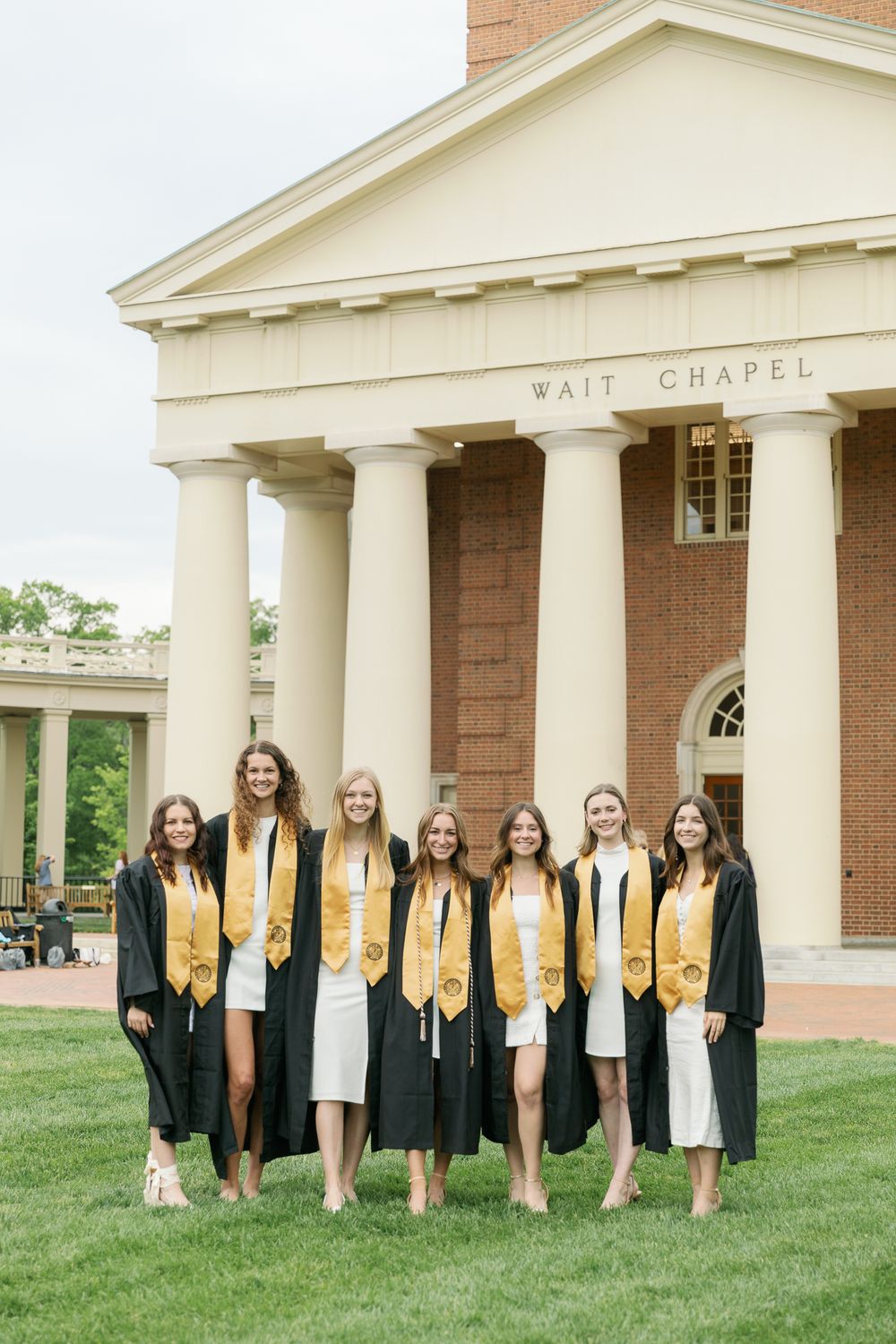 Graduates in black robes and gold stoles pose in front of Wait Chapel building.
