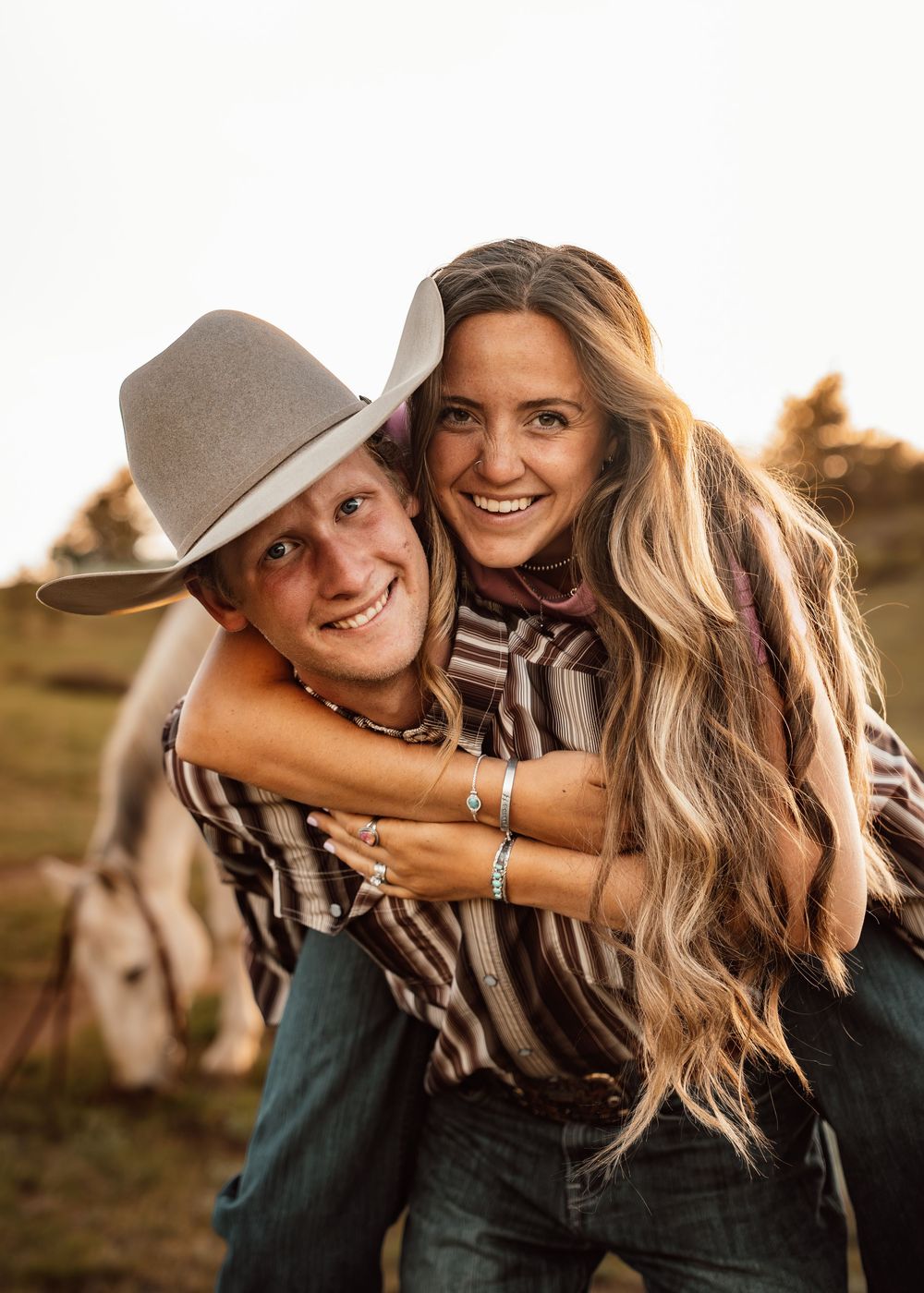 A smiling couple in western wear, man in cowboy hat giving woman a piggyback ride in a sunny field with a horse.