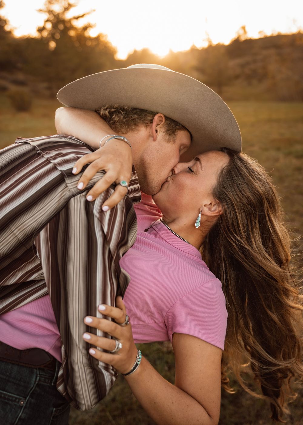 Couple sharing a romantic kiss at golden hour, woman in pink top and turquoise earrings, man in striped shirt and cowboy hat.