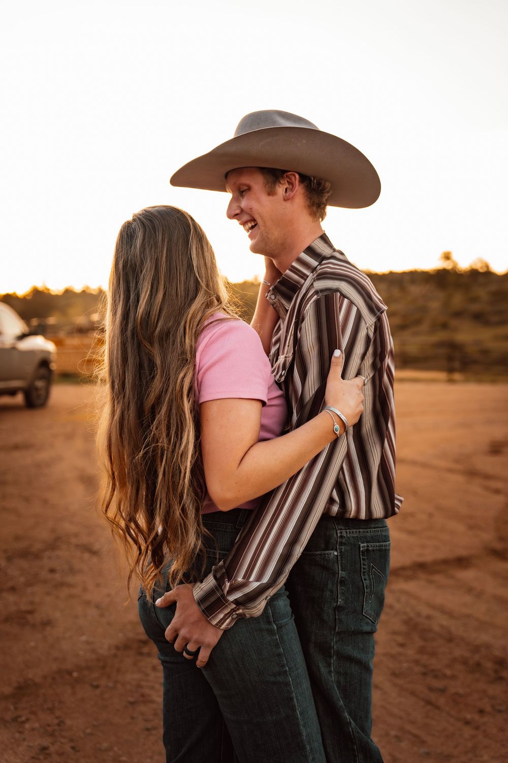 Couple embracing at golden hour, man in cowboy hat and striped shirt, woman in pink top and jeans, desert backdrop.