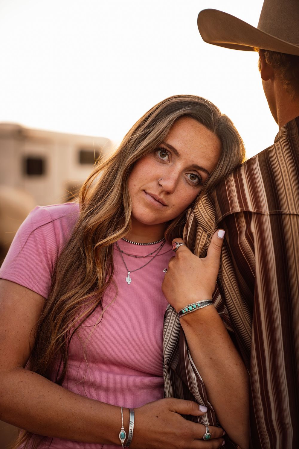 Young woman in pink tee with layered necklaces leans on man in cowboy hat and striped shirt outdoors.