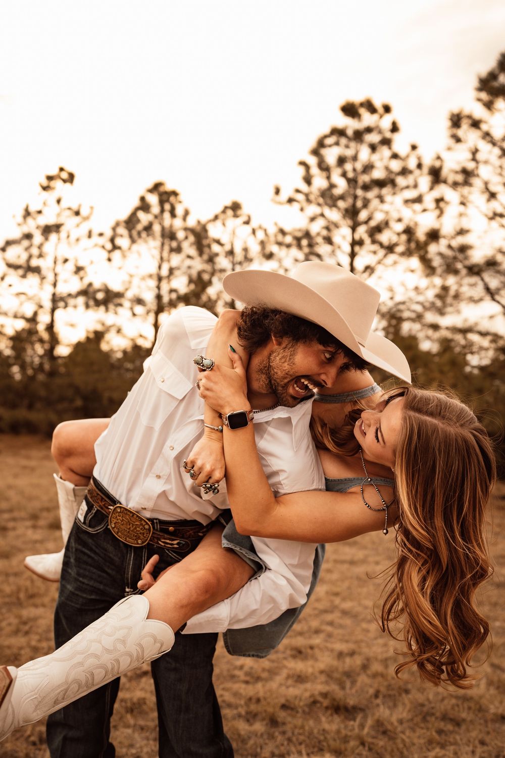 A joyful couple in western attire laughs outdoors, man lifting woman in white cowboy hat and boots at golden hour.