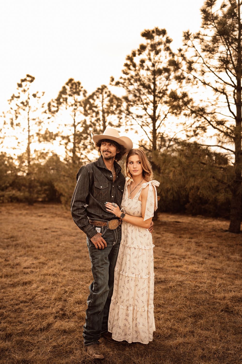 Couple in golden sunset field, man in cowboy hat and jeans, woman in floral maxi dress, embracing near pine trees.