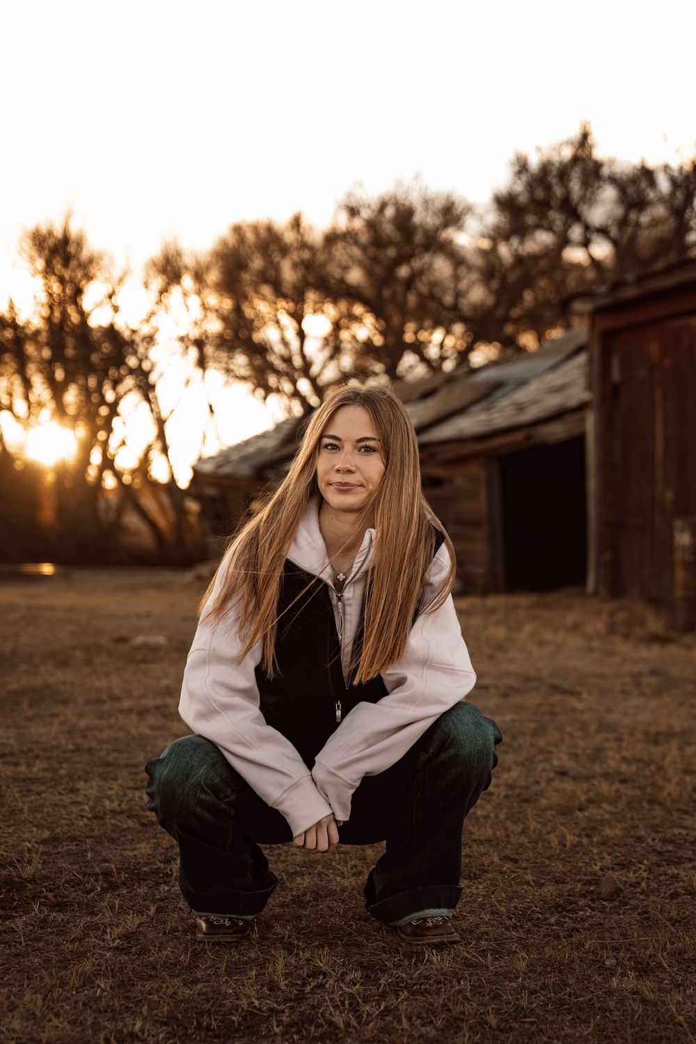 A person in casual attire sits in a grassy field at golden hour with a rustic barn setting in the background.