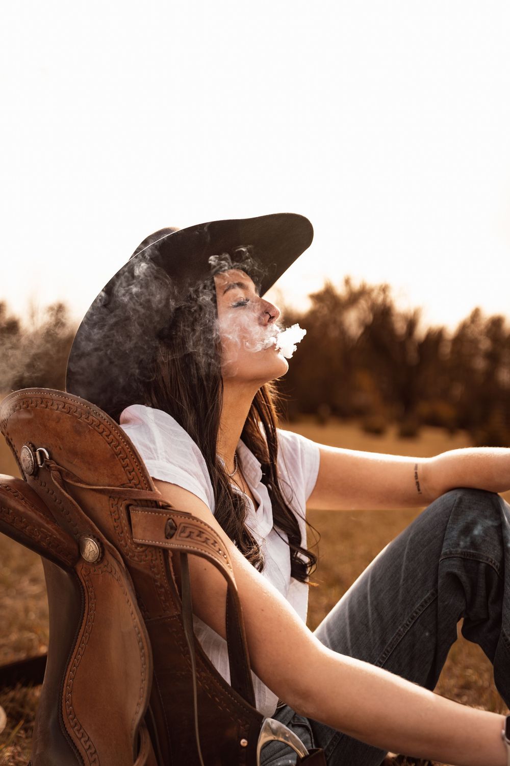 A young woman in a cowboy hat exhales smoke while relaxing outdoors in jeans and a white shirt at golden hour.