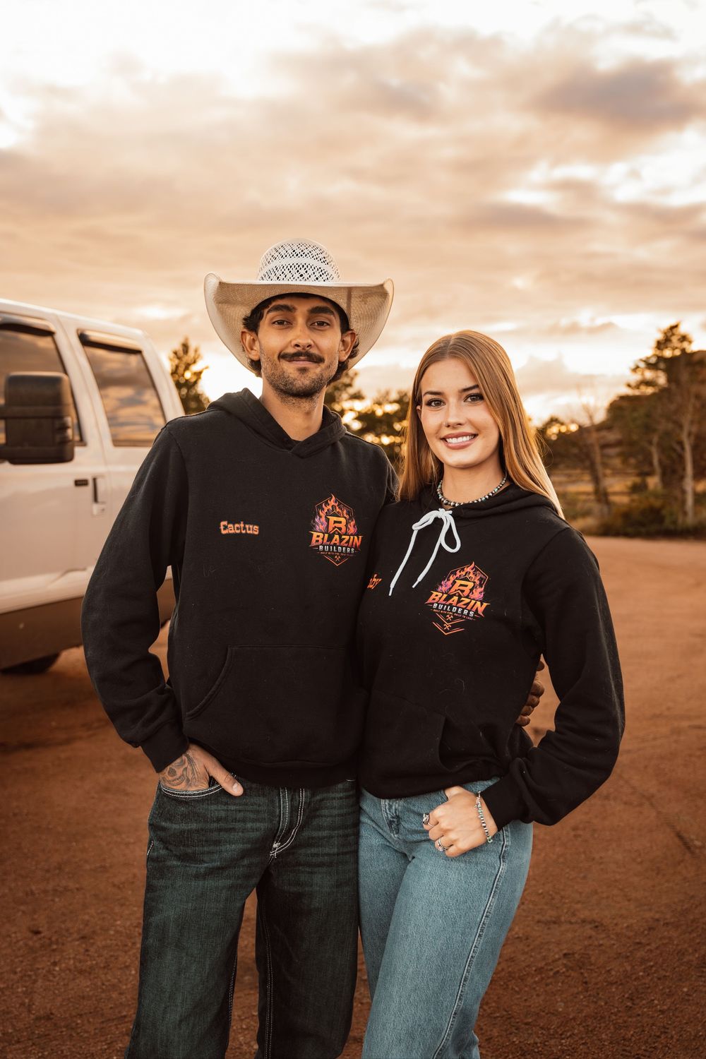 A man in a cowboy hat and woman wearing matching black Blazin hoodies pose together near a white truck at sunset.