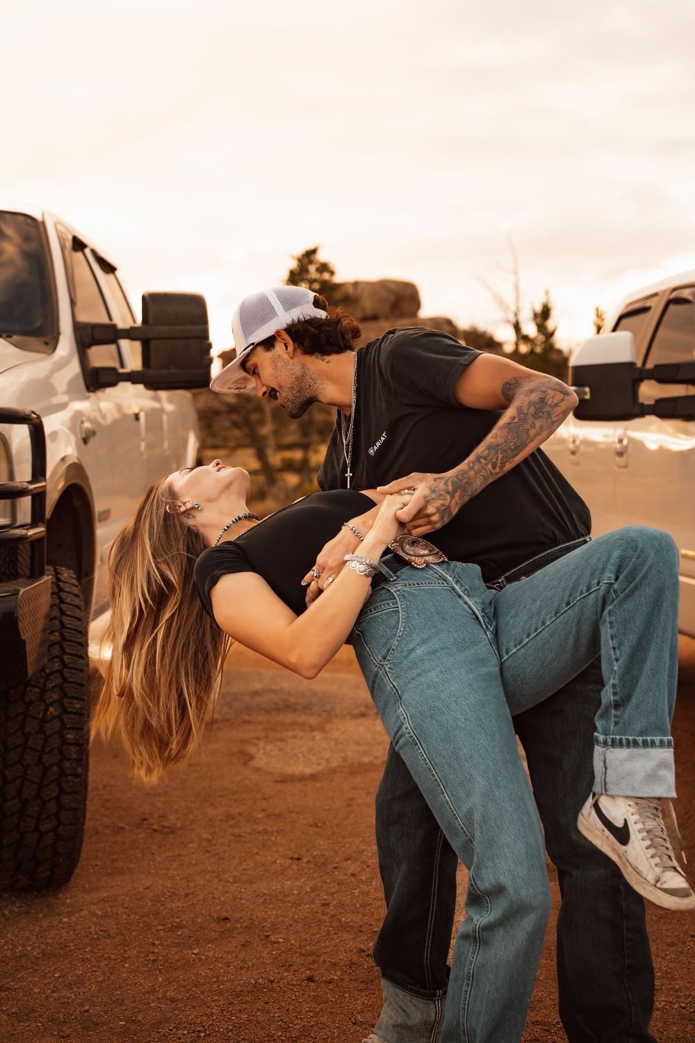 Couple sharing a romantic dip kiss between two white trucks on a red dirt road at sunset.