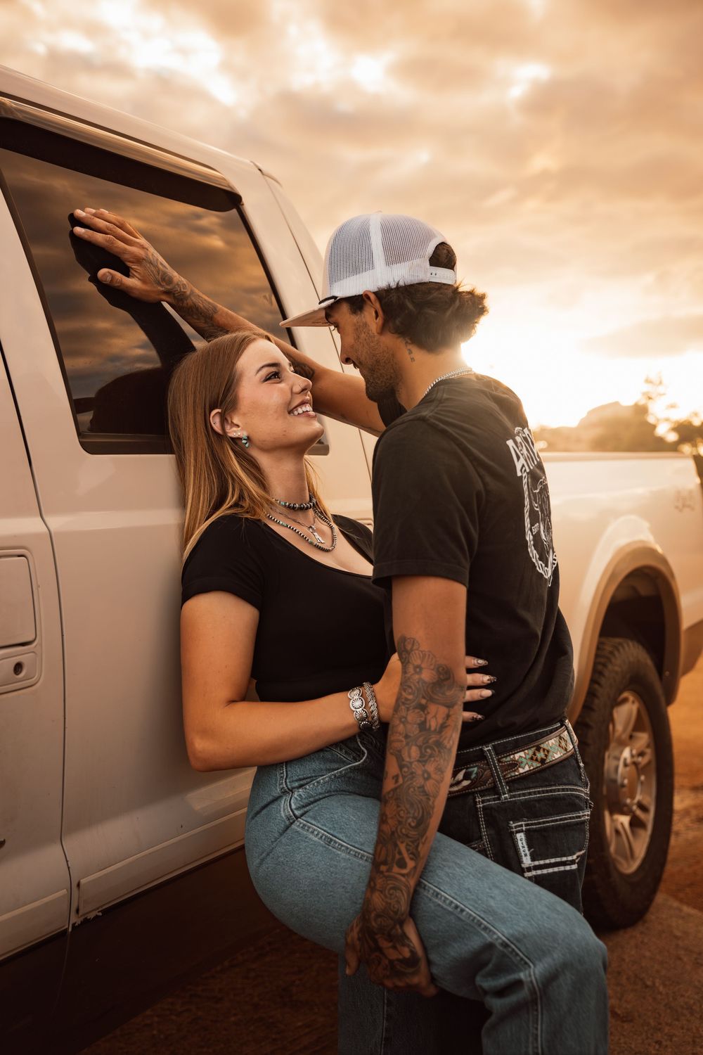 A tattooed couple shares a romantic moment leaning against a white pickup truck at golden sunset.
