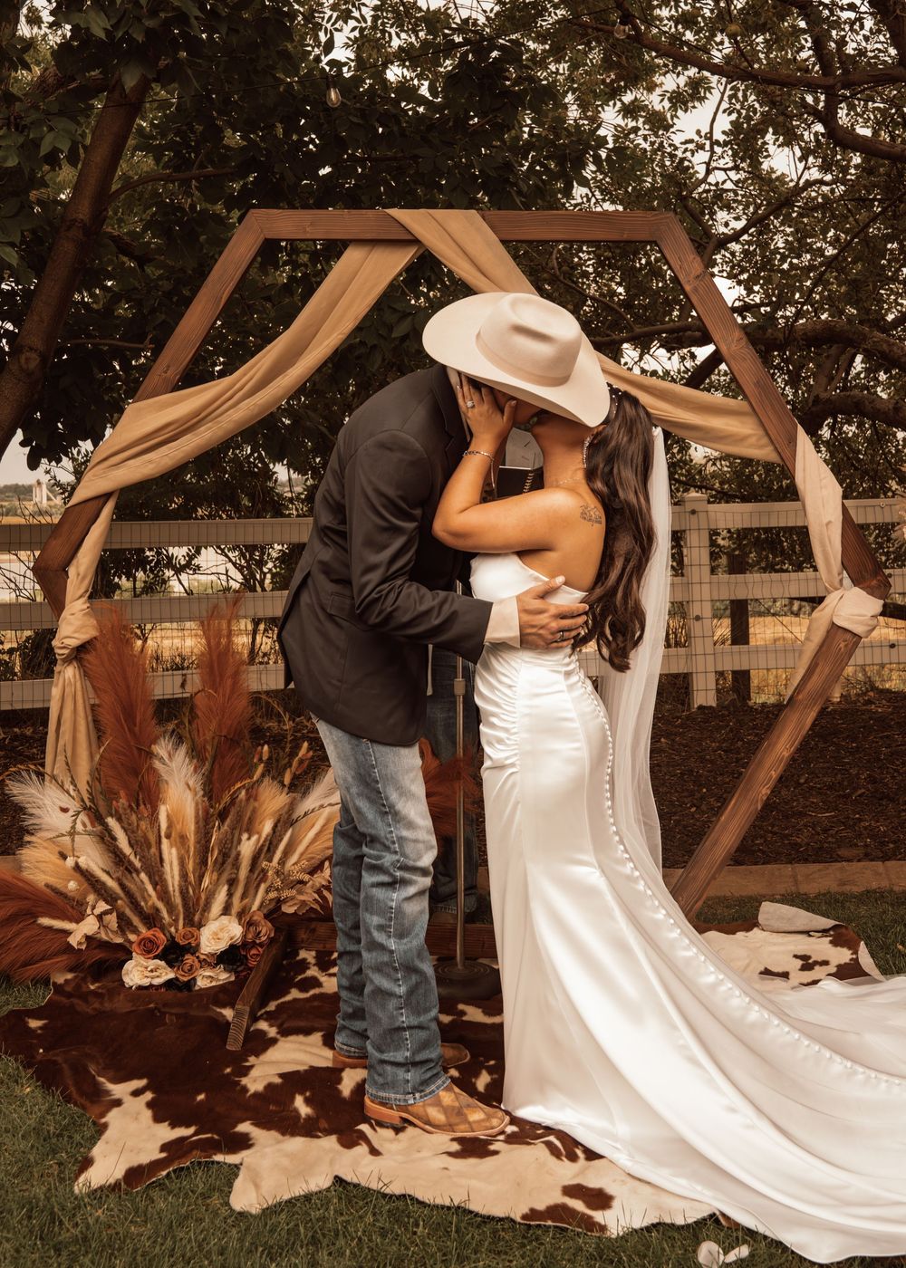 A romantic couple shares a dip kiss during their outdoor wedding ceremony under a rustic wooden arch.