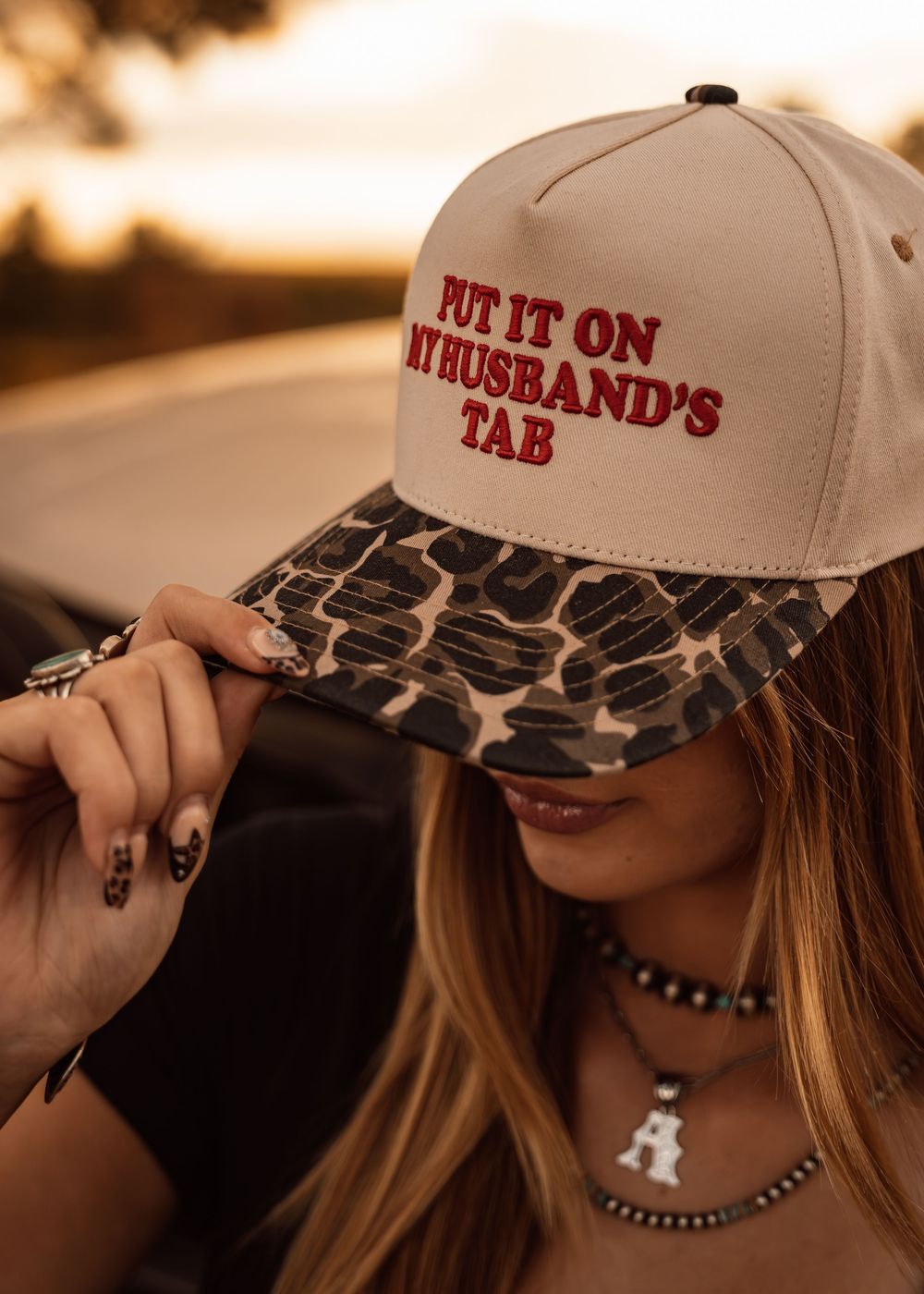 Woman wearing a beige snapback hat with leopard print brim reading "Put It On My Husband's Tab" at sunset.
