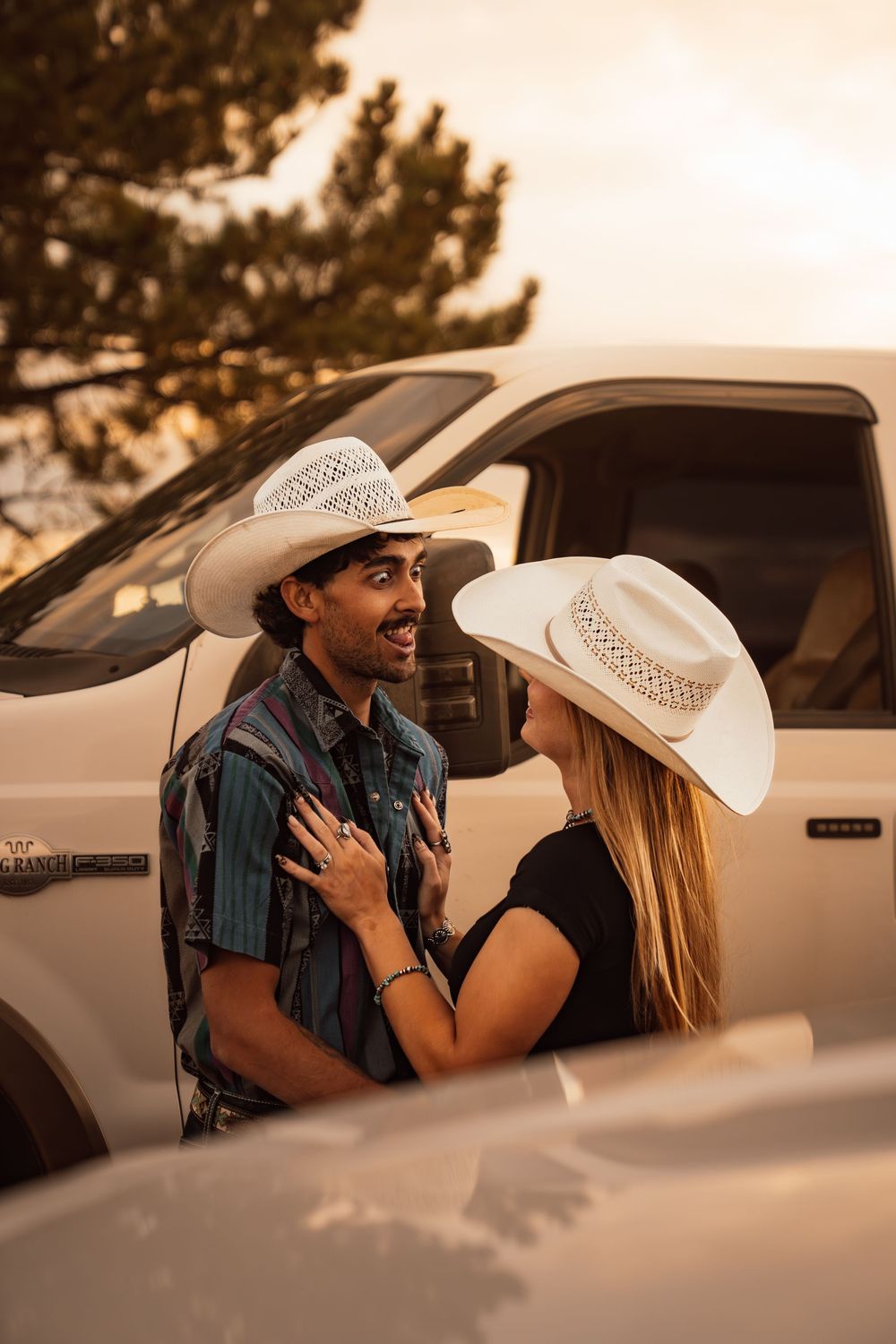 A couple in white cowboy hats share a romantic moment leaning against a white truck at golden hour sunset.