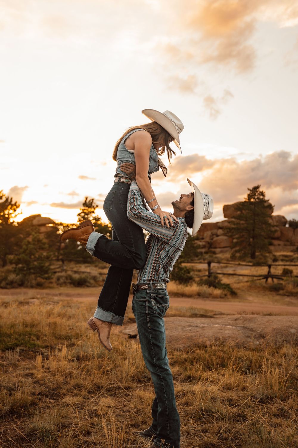A couple in cowboy hats shares a romantic lift at golden hour in a rustic field.