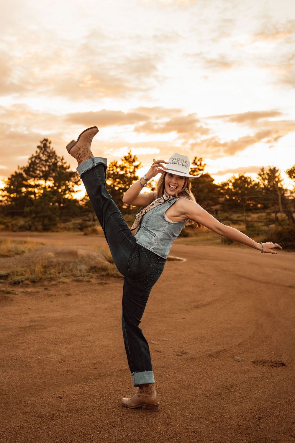 Woman in cowboy hat and boots kicking leg up on dirt road at sunset.