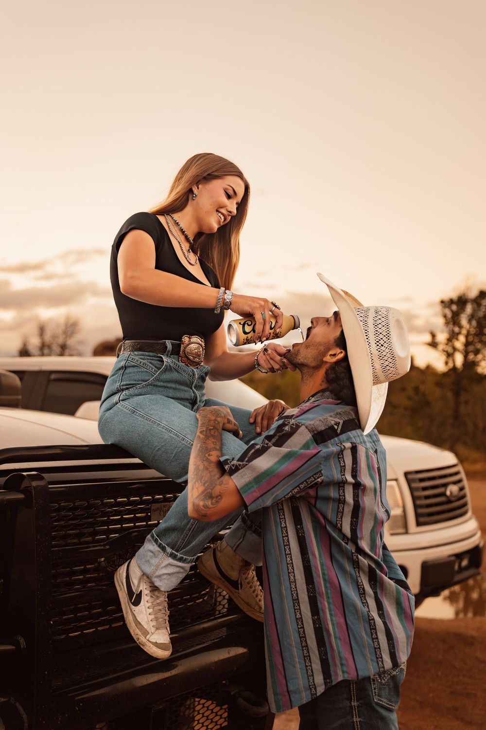 A couple in Western attire share a drink by a pickup truck at sunset, with the woman sitting on the hood.