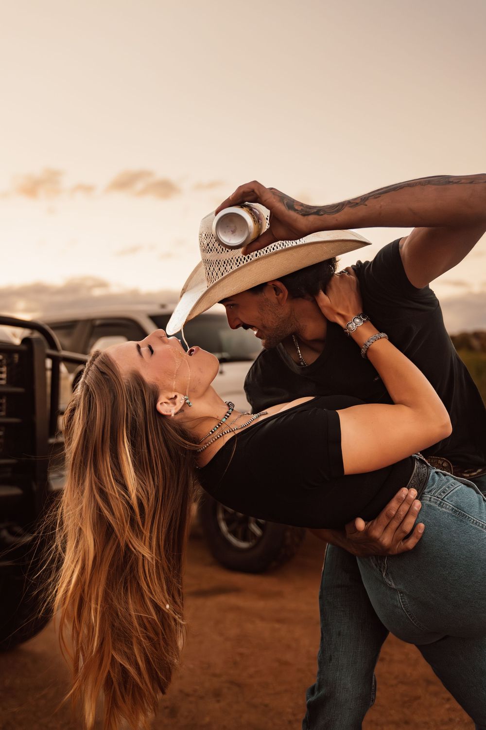 A couple shares a romantic dip dance at sunset near a truck, the man tipping his cowboy hat over her.