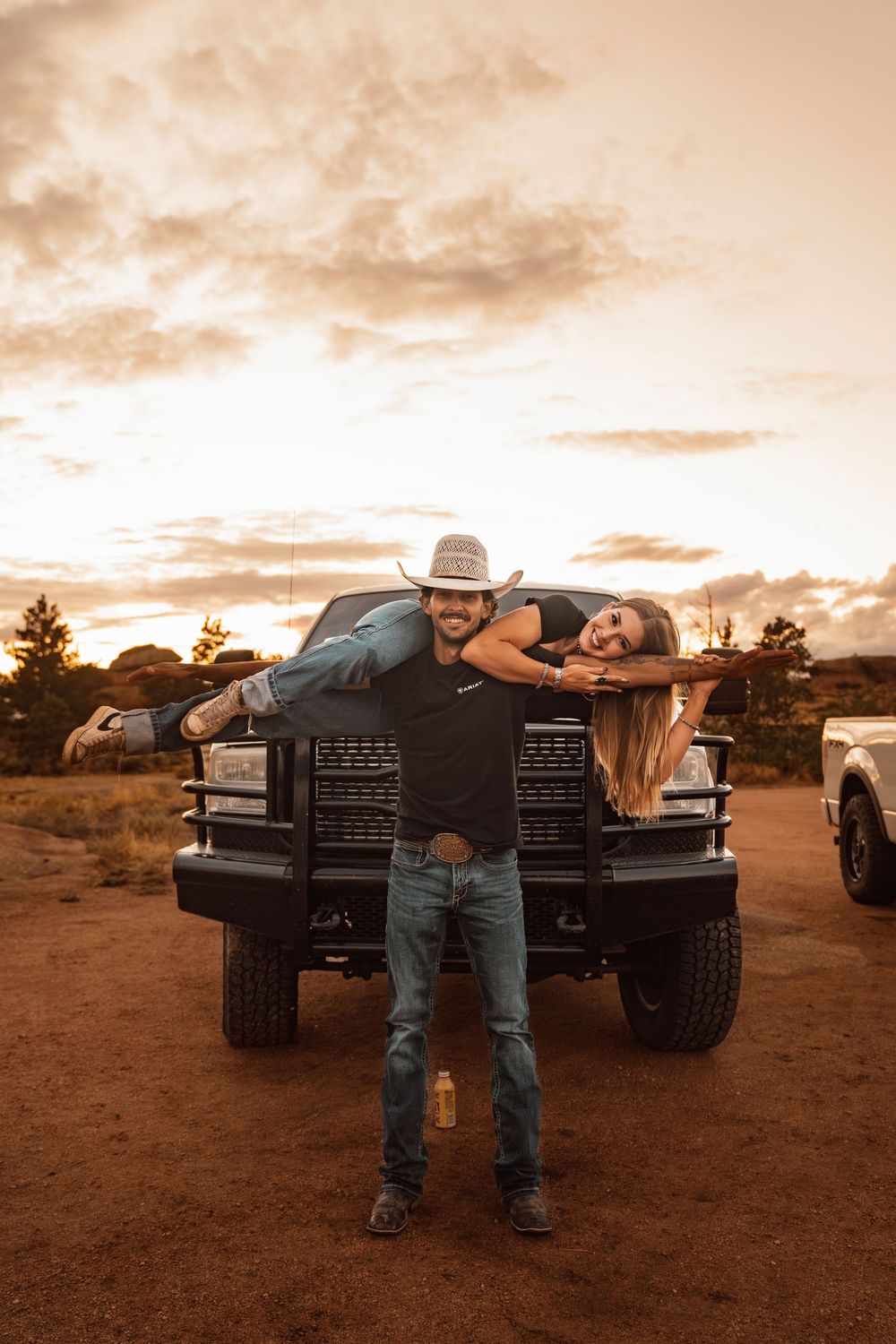 A man in a cowboy hat carries a woman on his shoulder in front of a black truck at sunset on a dirt road.