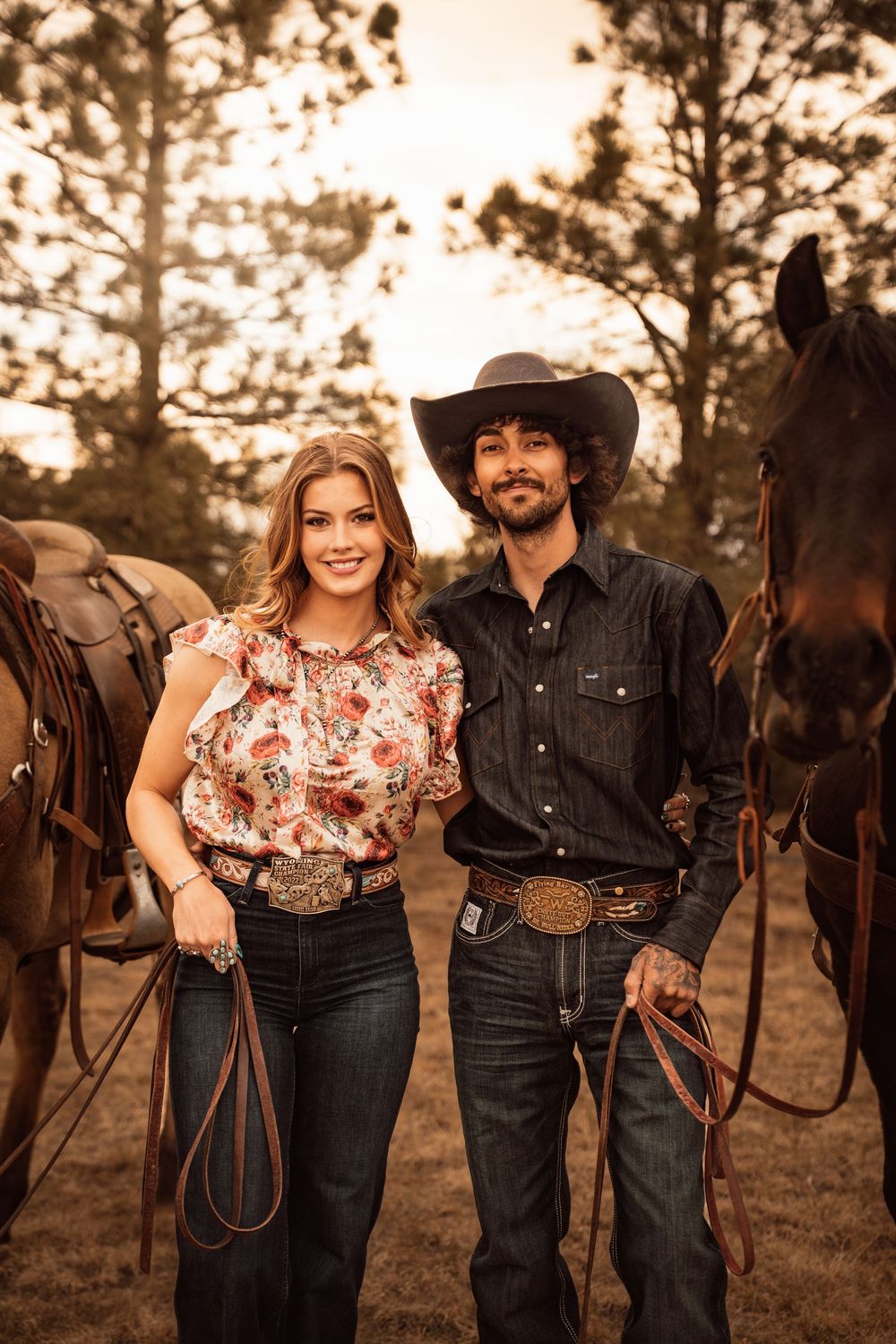 A smiling couple in western attire with belt buckles and cowboy hat stands beside saddled horses at golden hour.
