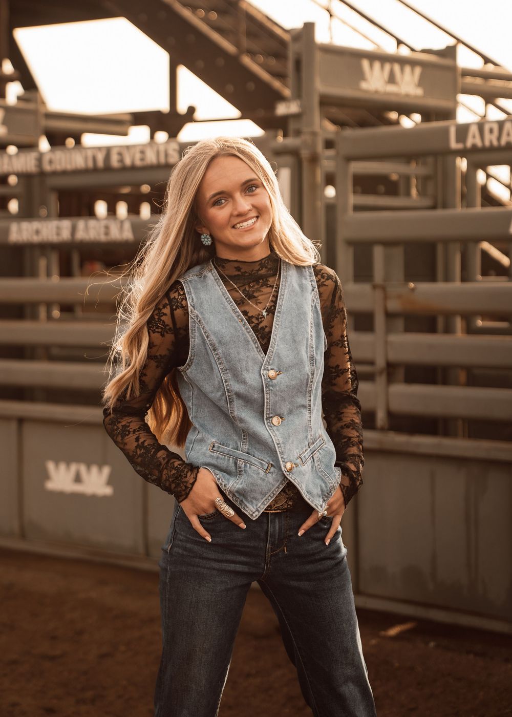 Young woman in denim vest and lace top smiling at rodeo arena, Archer Arena county events backdrop.