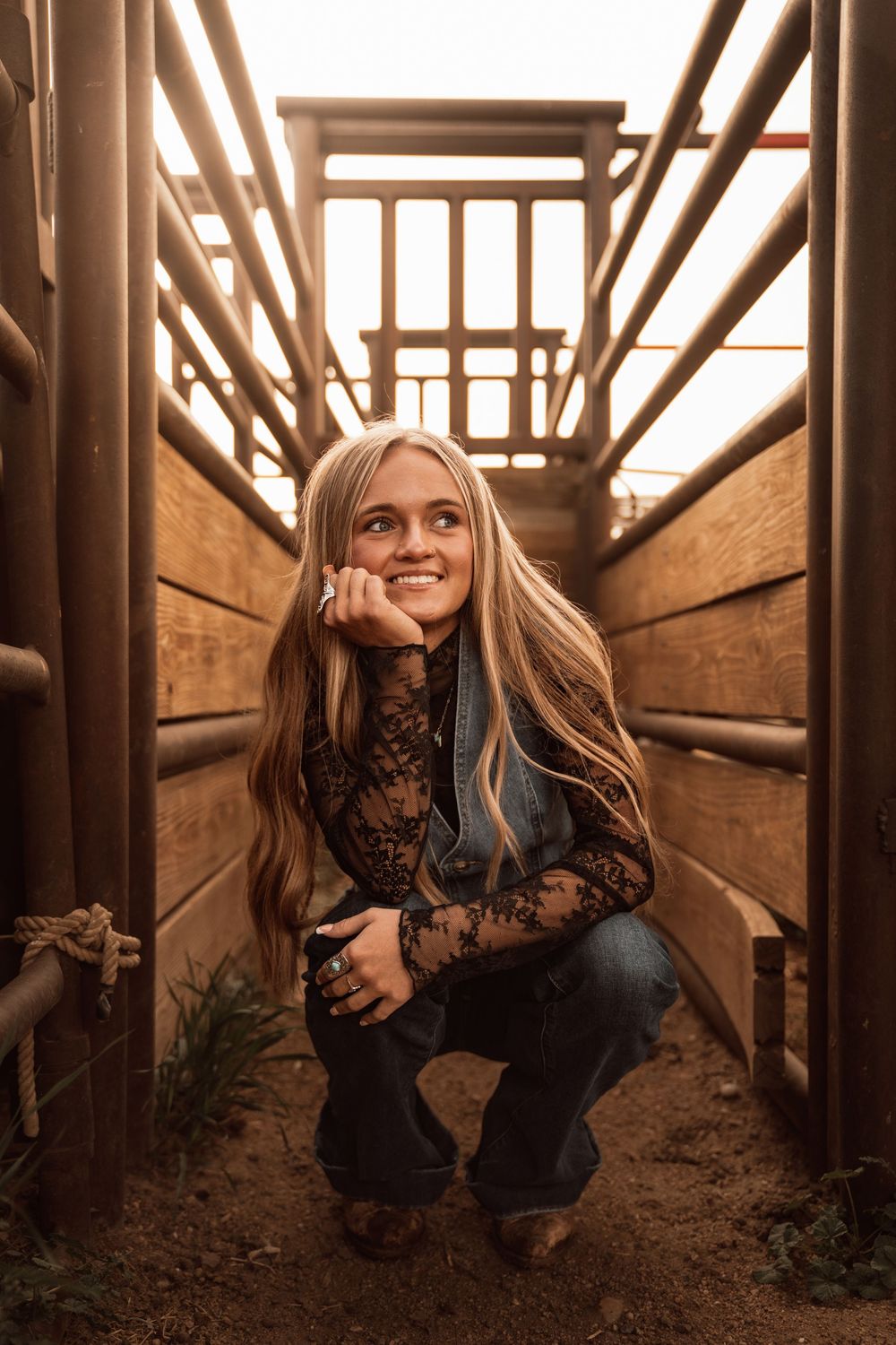 Smiling blonde girl crouching in rustic wooden cattle chute, wearing lace top and jeans, bathed in warm golden light.