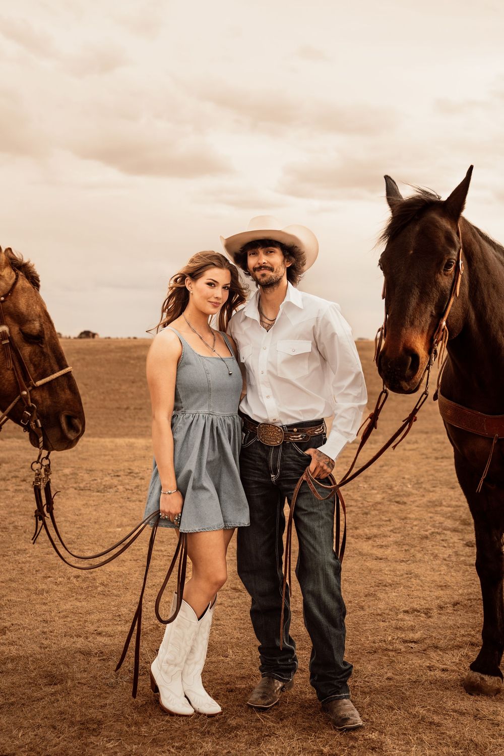 A couple in western attire poses with two brown horses on a ranch under a dramatic cloudy sky.