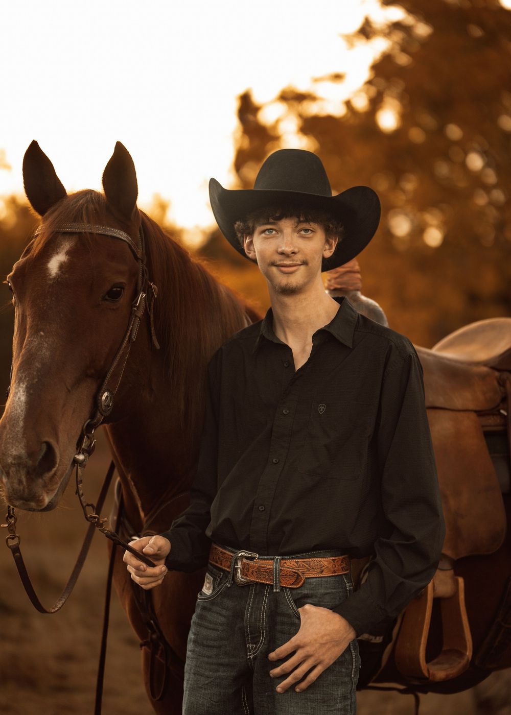 Western-styled person in black shirt and cowboy hat standing with brown horse at sunset.