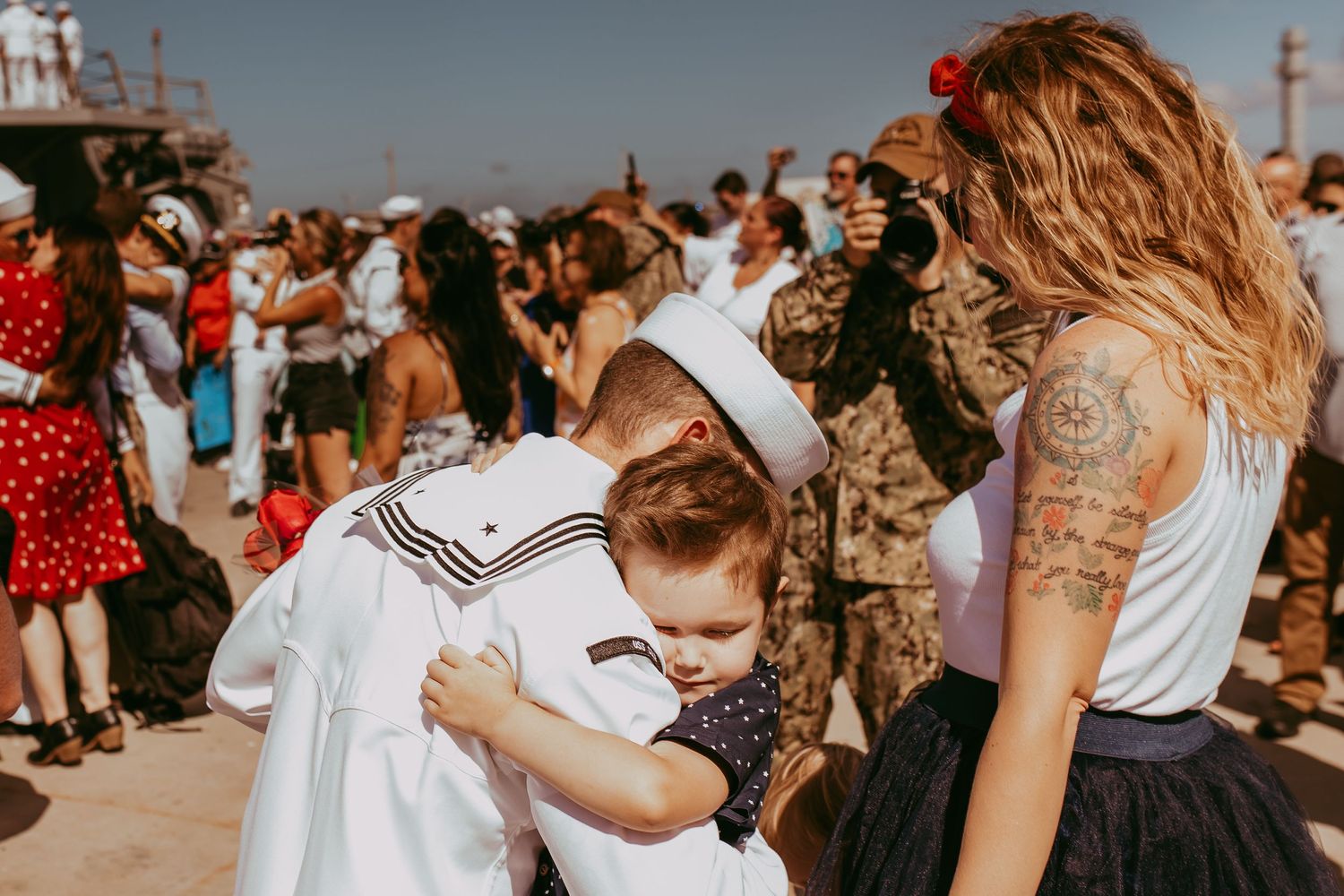 Emotional reunion scene at military homecoming with sailor in dress whites surrounded by crowd on sunny day.