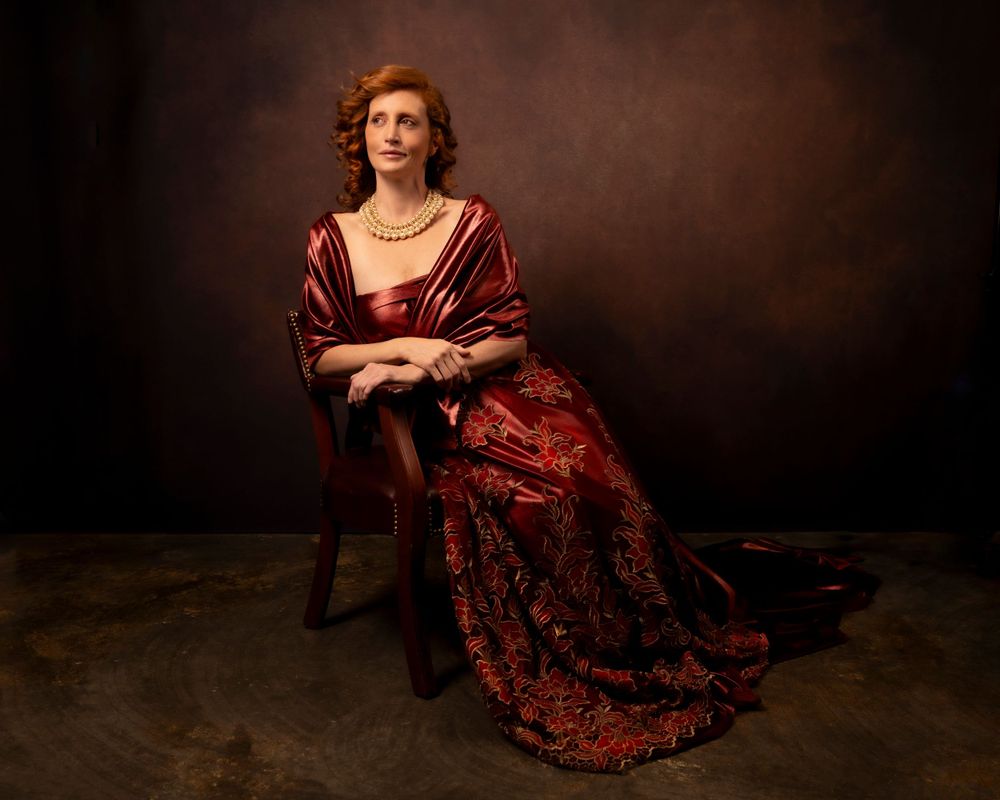 Classical portrait in burgundy Renaissance-style dress seated against dark background.