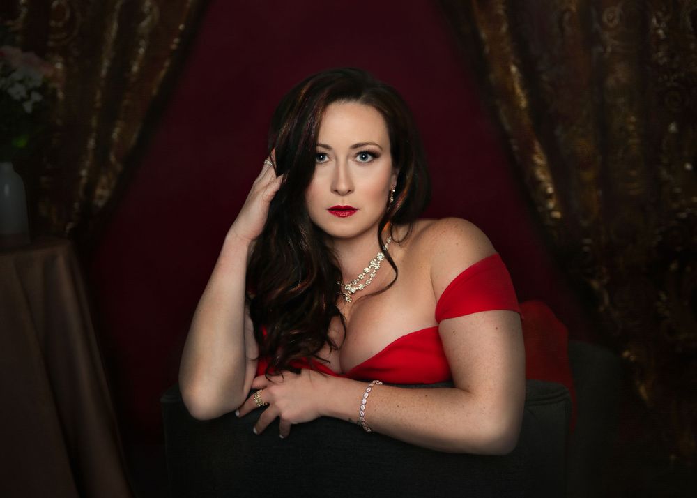 Dramatic portrait in a red dress against dark curtains with moody lighting and elegant jewelry.