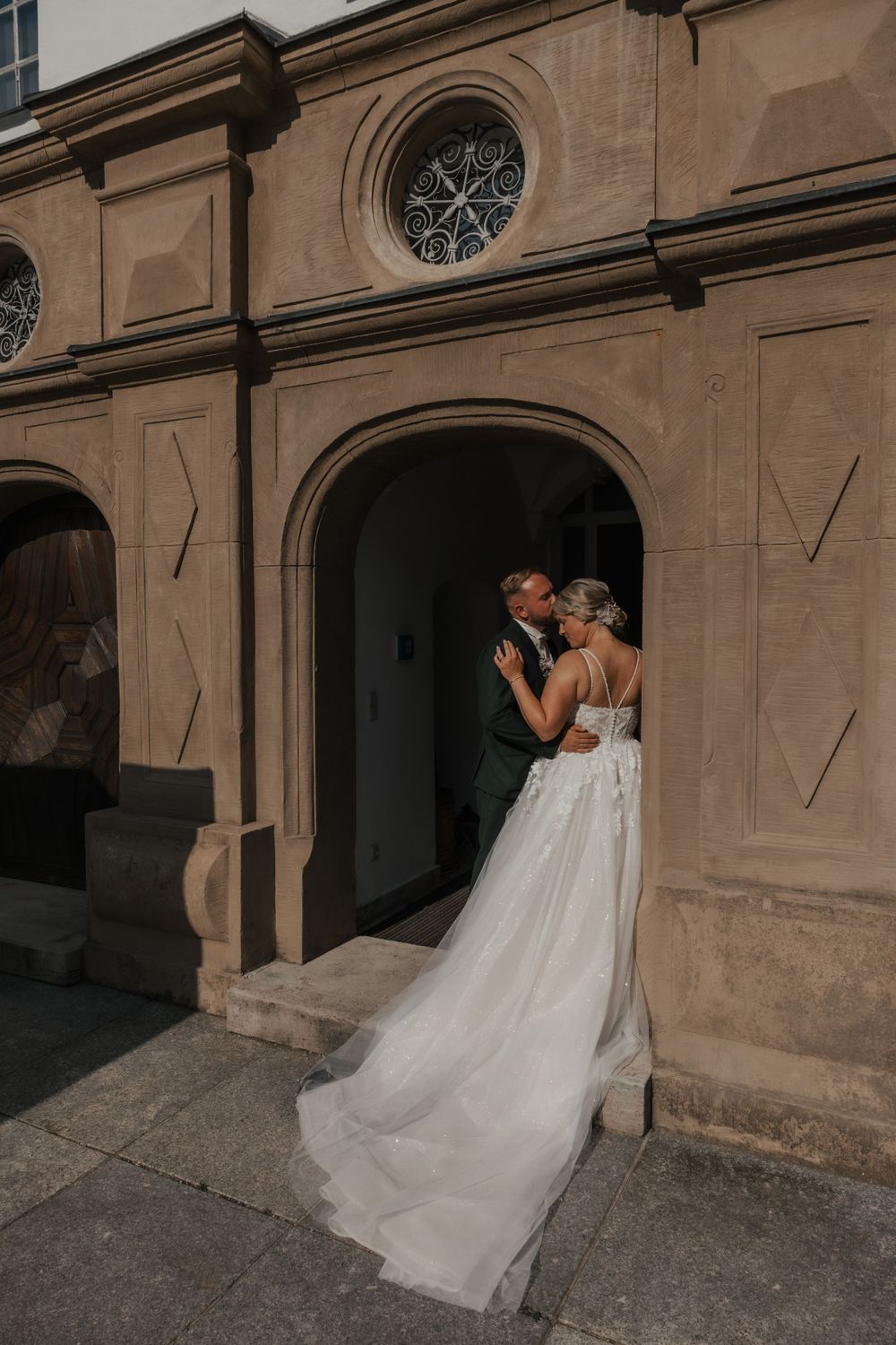 Wedding couple embracing in stone archway of historic building as dress train flows behind.
