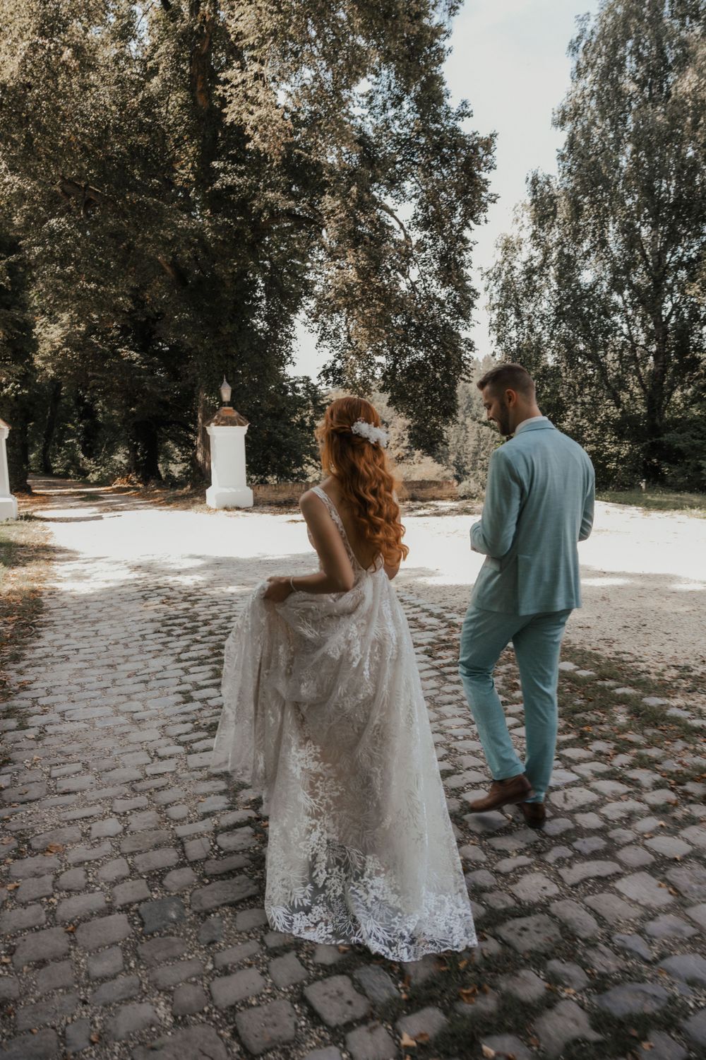 Wedding couple walking along a white wall and cobblestone path in an outdoor garden setting.