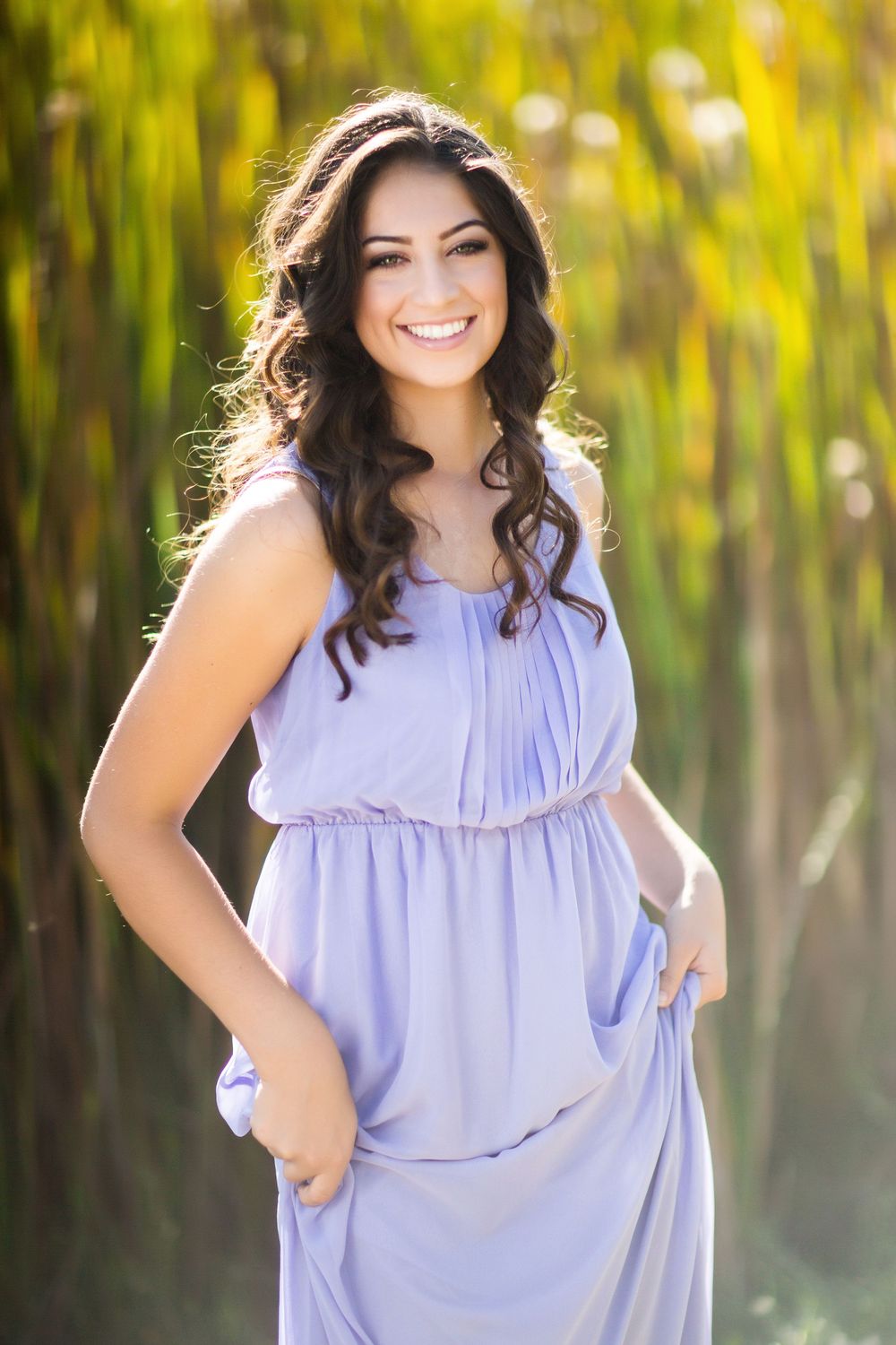A woman in a flowing lavender dress poses outdoors against a blurred yellow and green natural background.