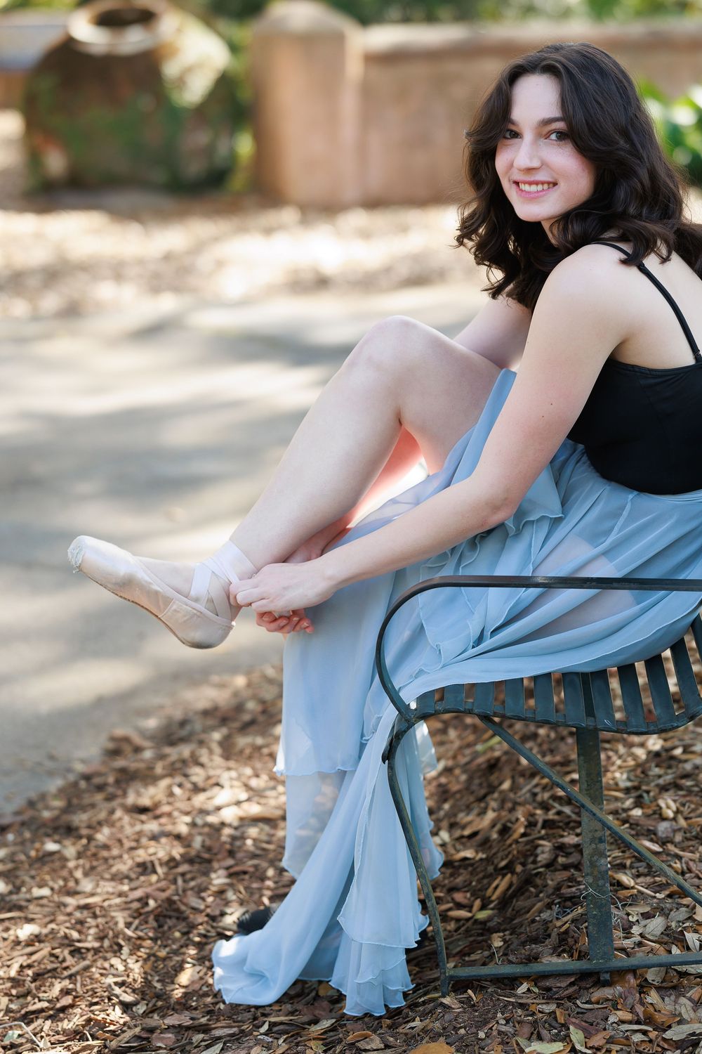 A ballet dancer in a blue skirt adjusts her pointe shoes while sitting on a park bench in dappled sunlight.