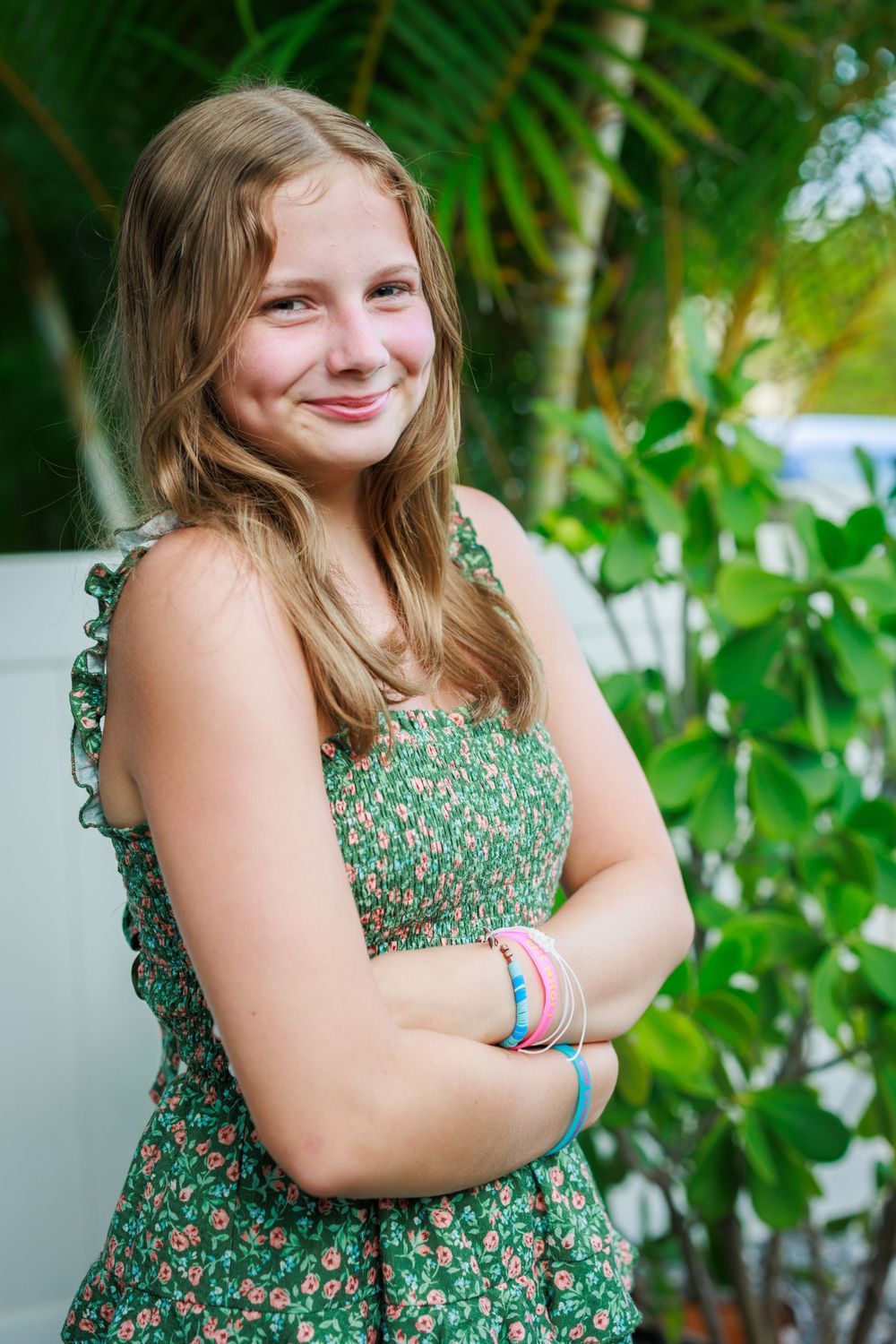 Person in a floral summer dress poses casually with crossed arms against tropical foliage.
