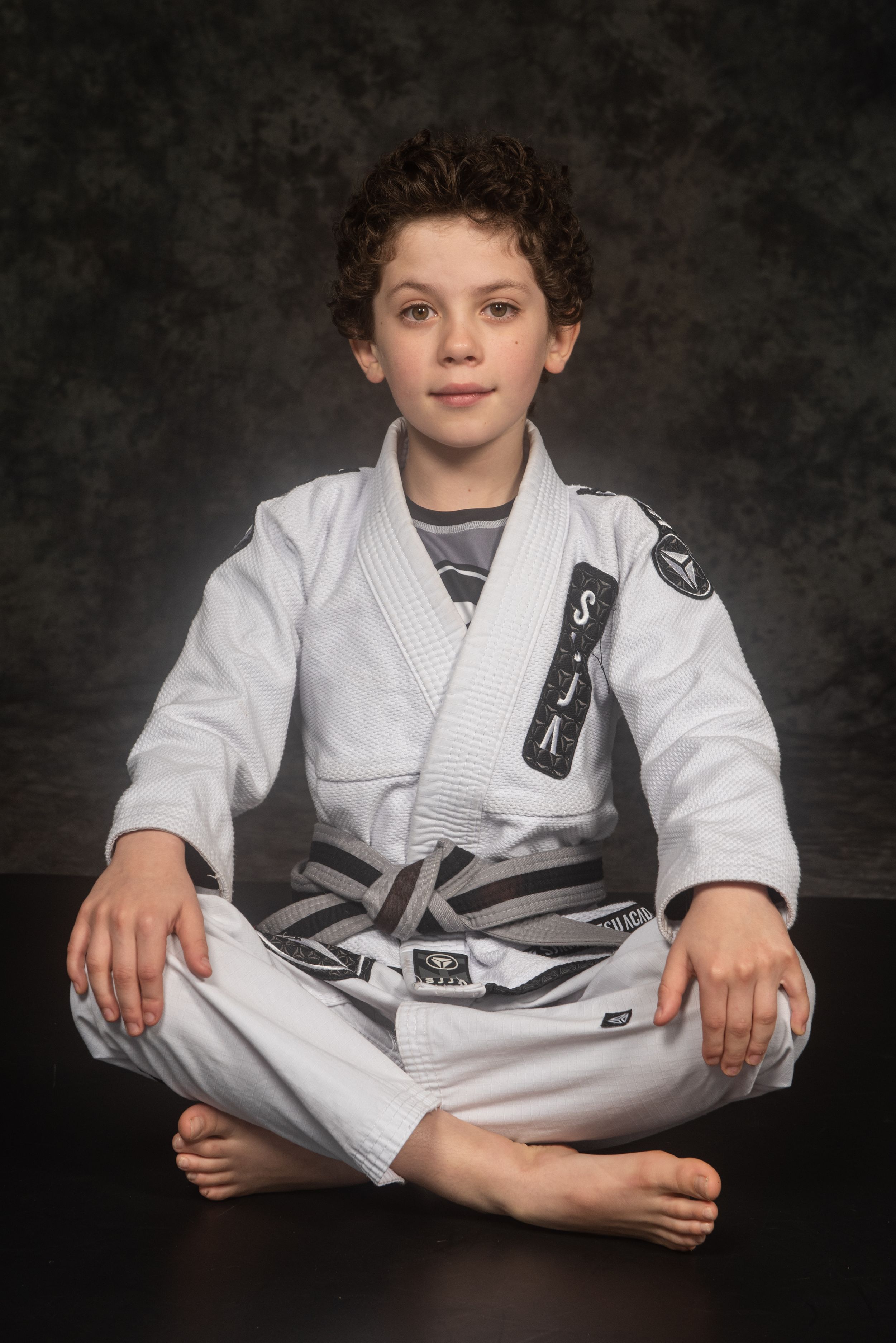 A young martial arts student sits cross-legged in a white gi uniform against a dark background.