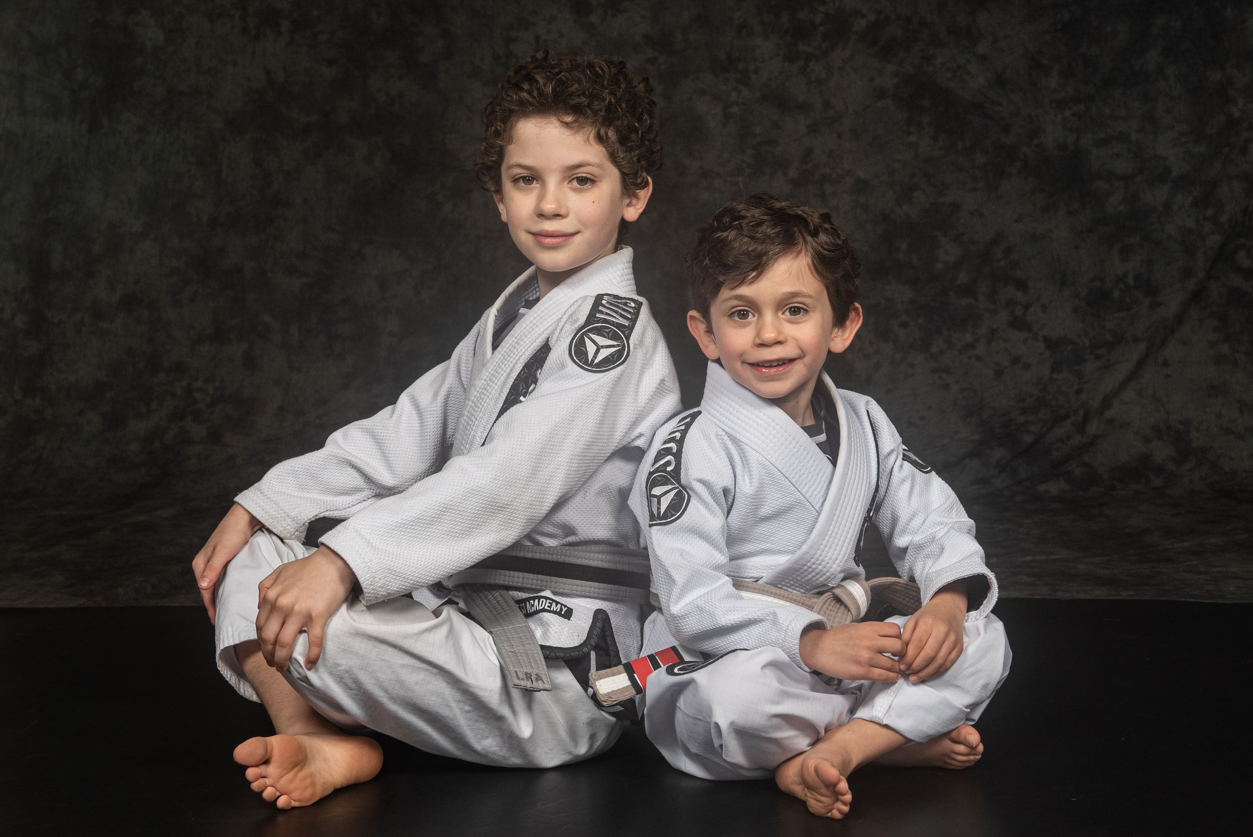 Two people in white martial arts gis pose playfully together on a dark backdrop.