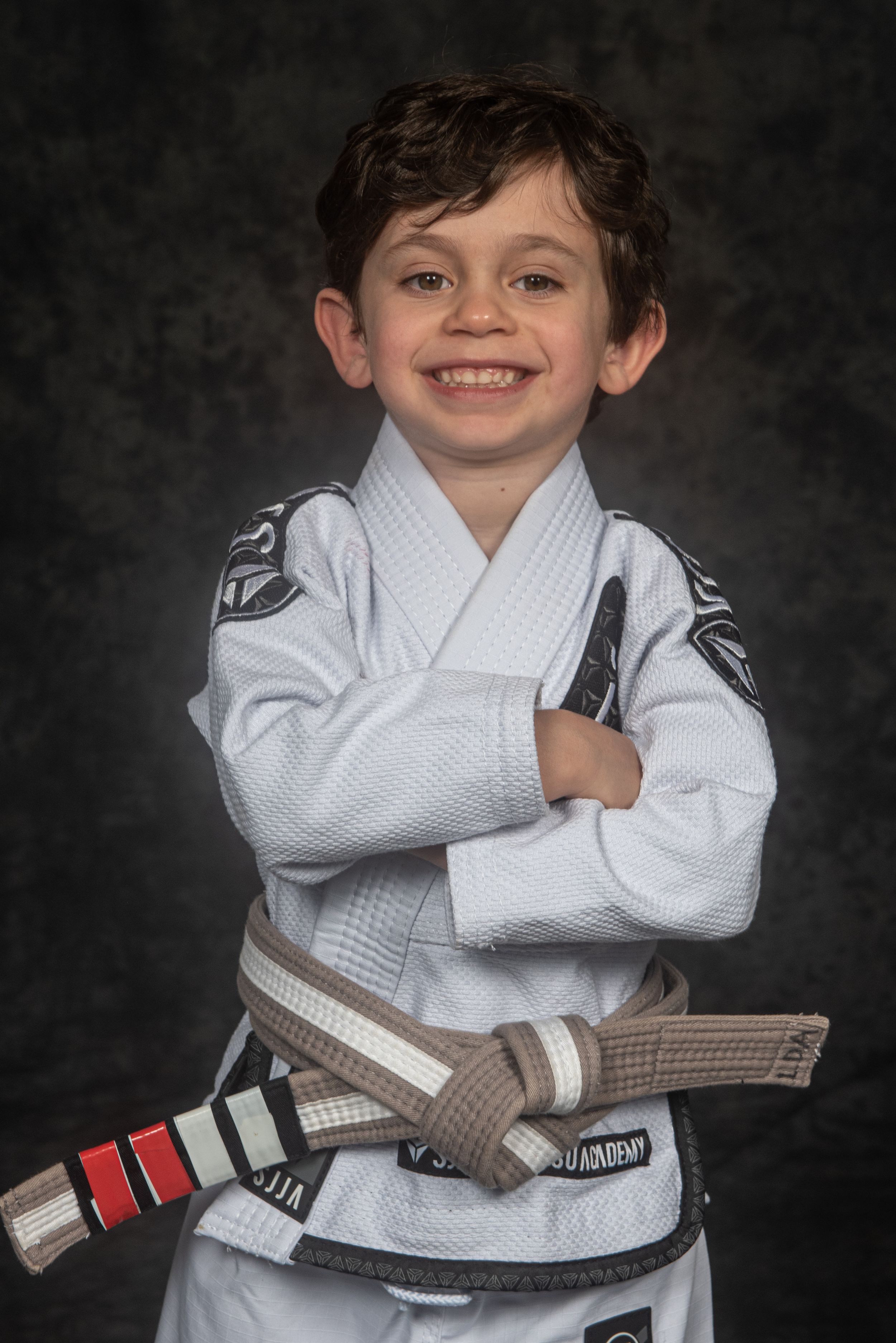 A martial arts student wearing a white gi and brown belt poses confidently against a dark background.