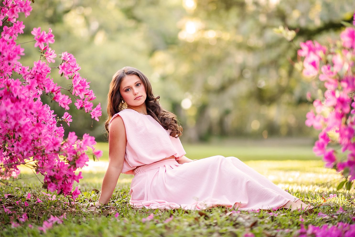 Young woman in pink dress sitting gracefully among bright pink bougainvillea flowers outdoors.
