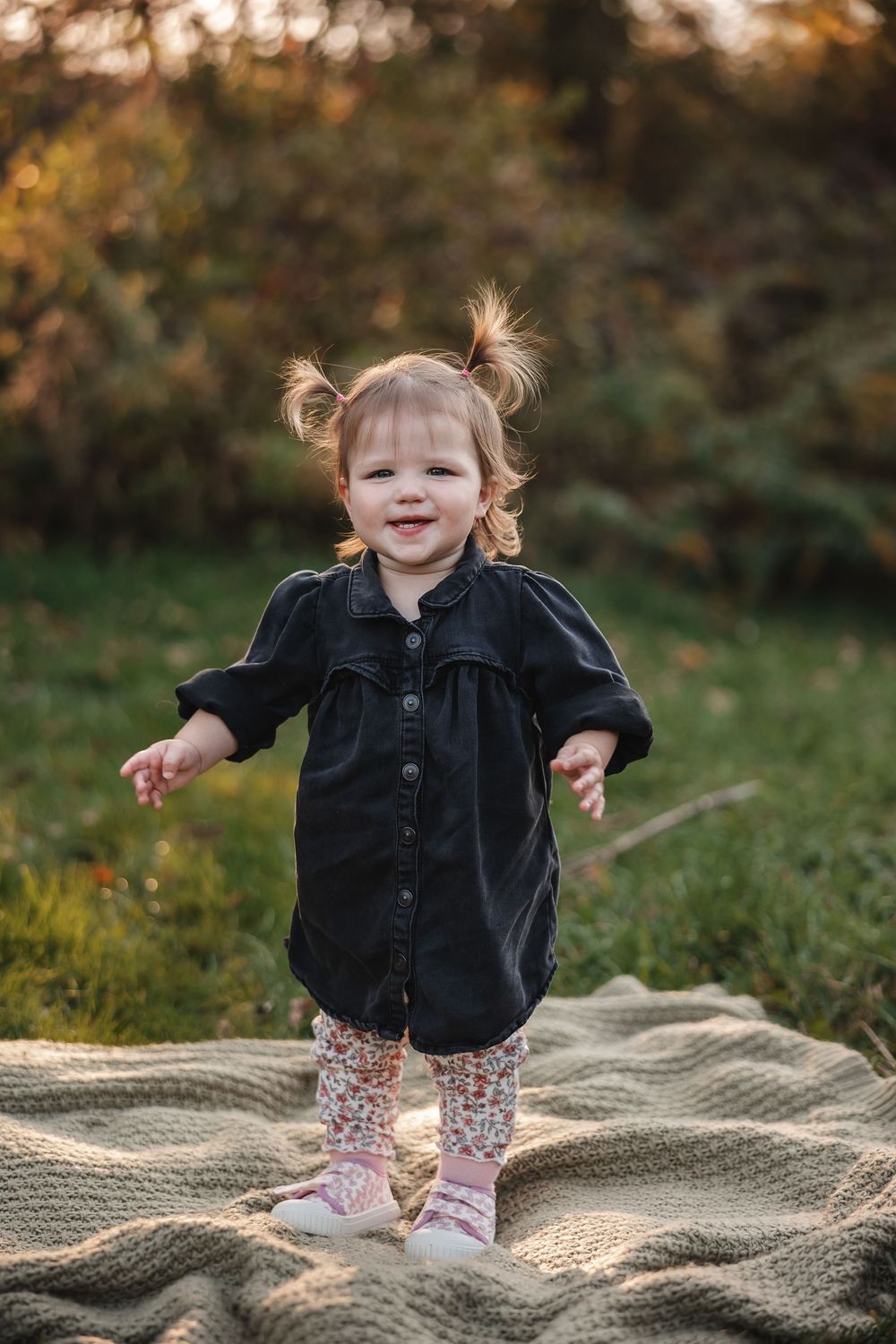 A small child wearing a black jacket and floral pants plays joyfully on a textured blanket in an autumn outdoor setting.