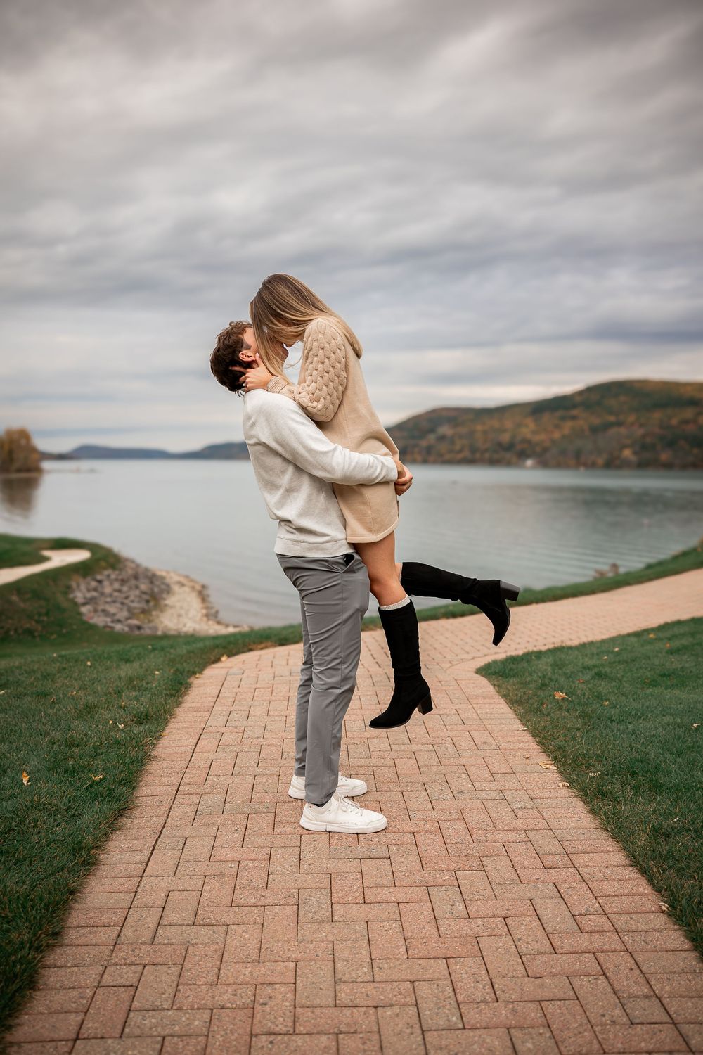 A joyful celebration moment captured on a lakeside brick pathway with dramatic cloudy skies and autumn colors.