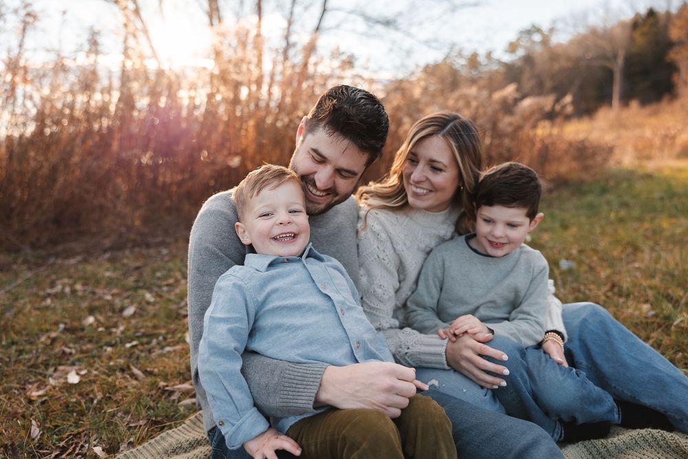 Family wearing coordinated blue outfits sits together in a grassy field during golden hour sunset.