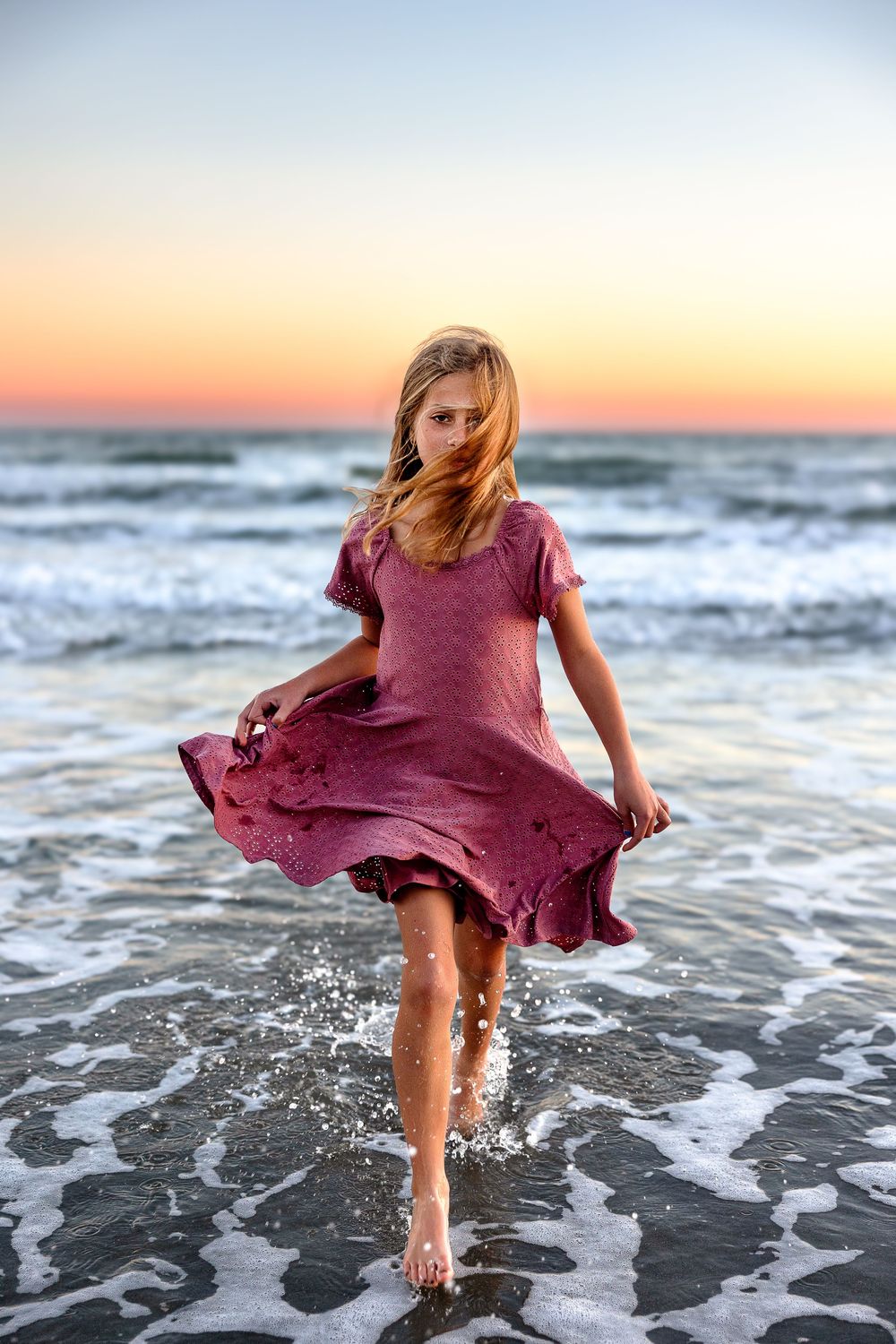 A person in a flowing burgundy dress splashes through ocean waves at sunset on the beach.